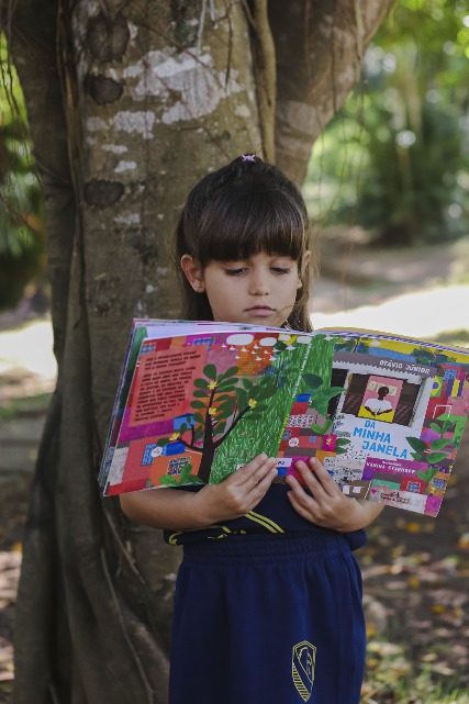 Criança da Educação Infantil lendo um livro em espaço externo da escola