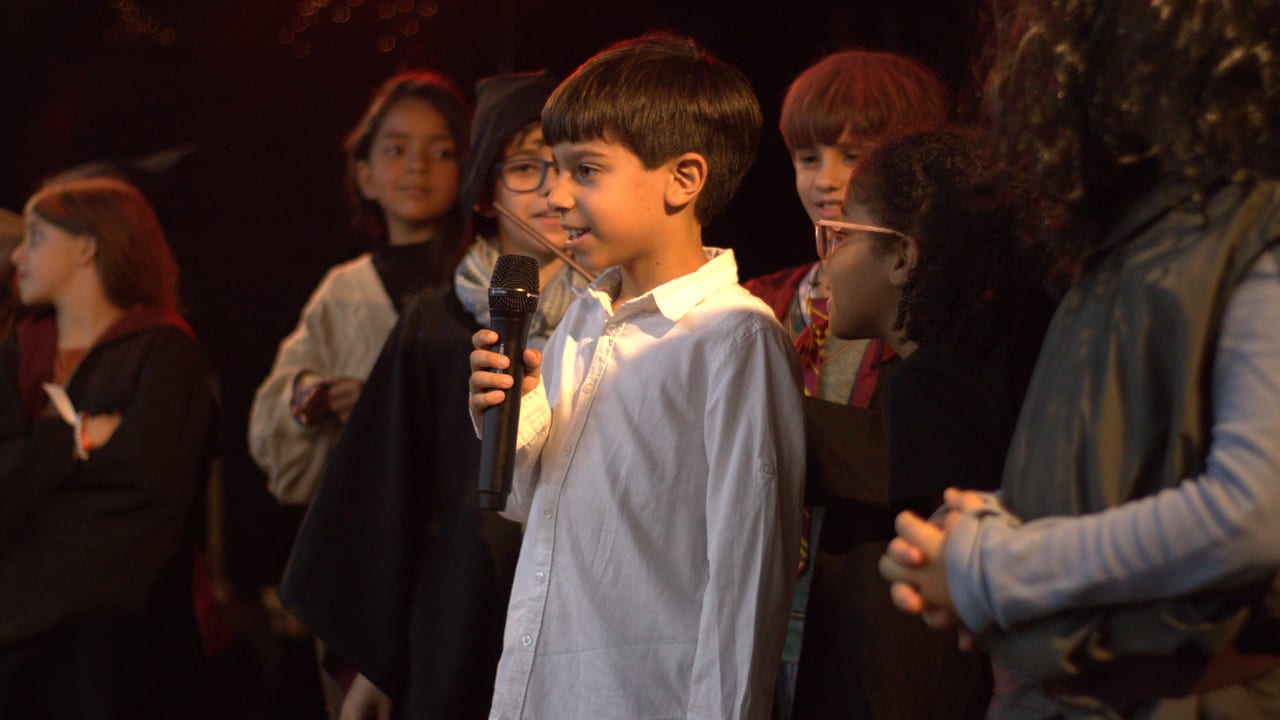 Aluno falando ao microfone em apresentação de teatro em inglês, com colegas fantasiados ao fundo
