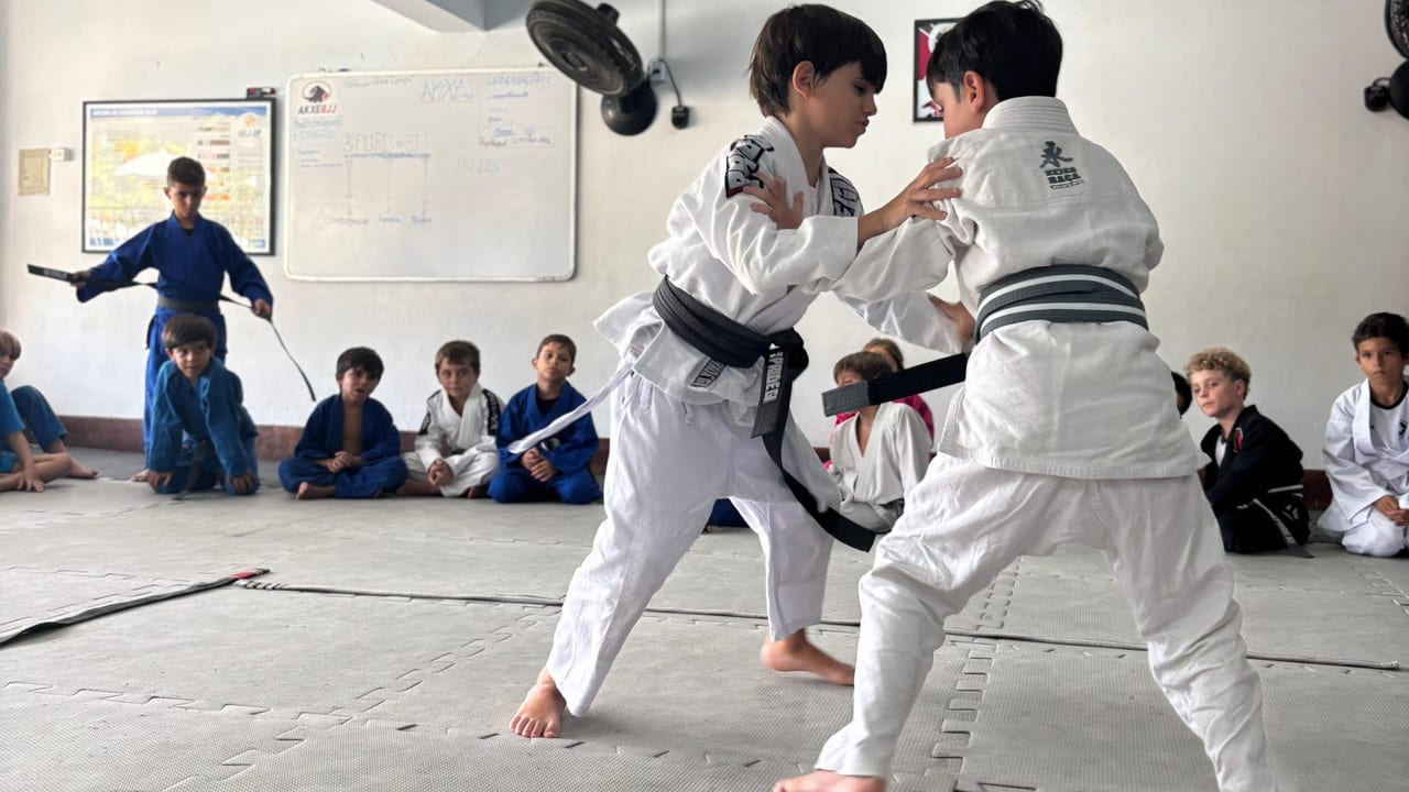 Estudantes de quimono treinando Jiu-Jítsu no tatame enquanto a turma observa